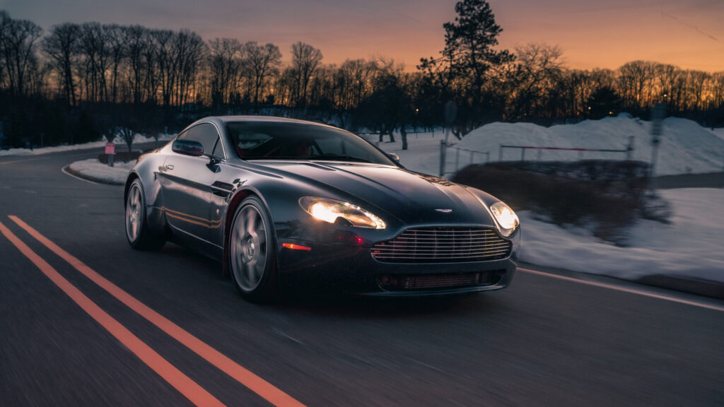 Aston Martin V8 Vantage running on a snow-lined road at sunset with headlights lit