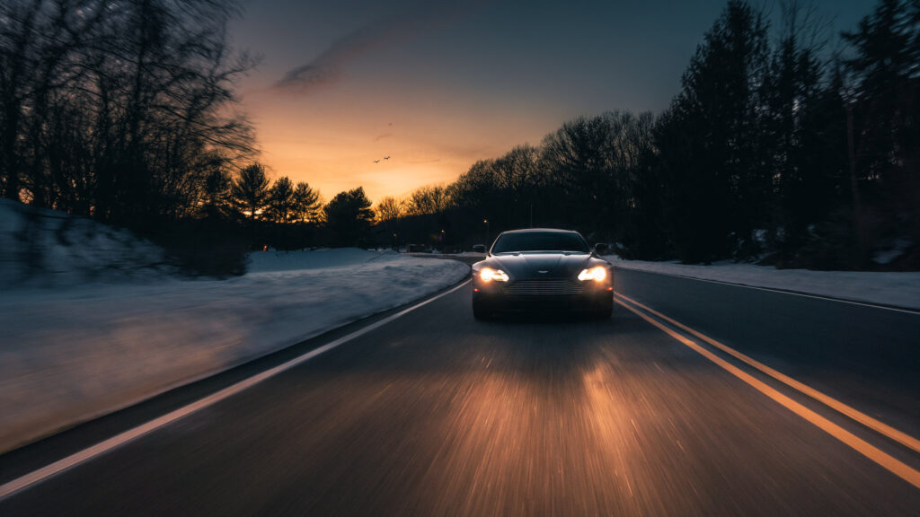 Aston Martin V8 Vantage head-on at dusk, headlights lit, sunset behind a tree-lined road
