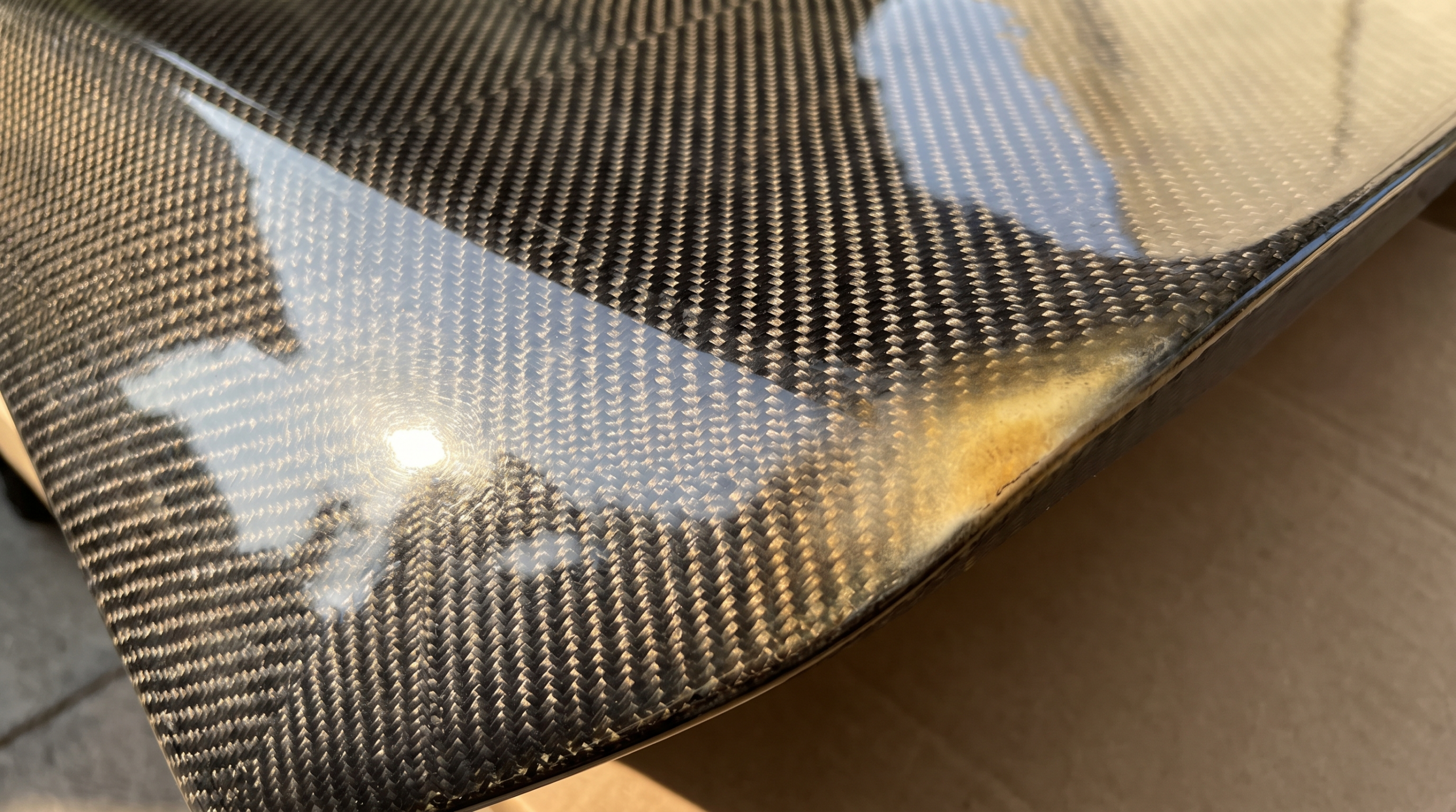 Exposed twill-weave carbon fibre panel under hard light, with slight yellowing at one edge