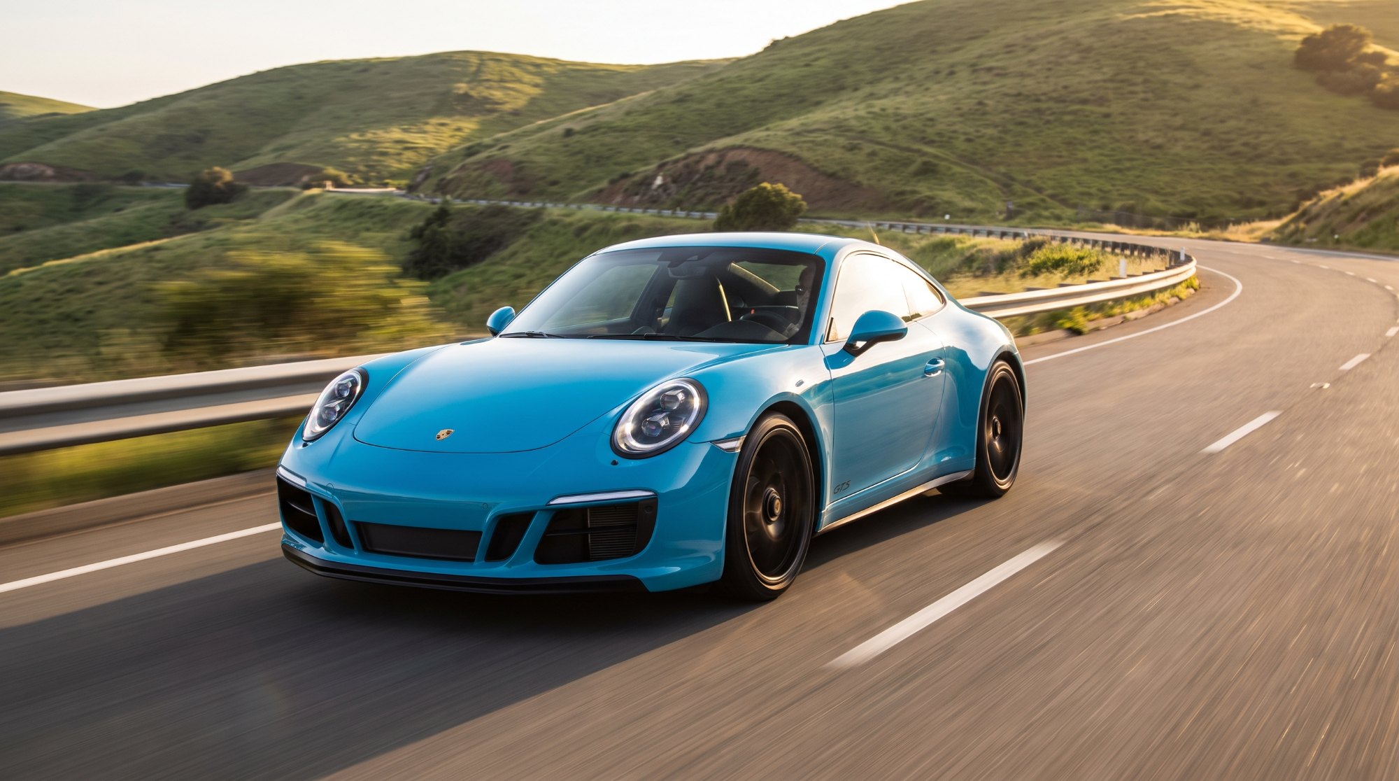 Miami Blue Porsche 911 Carrera GTS of the 991.2 generation cornering on a winding road through green hills