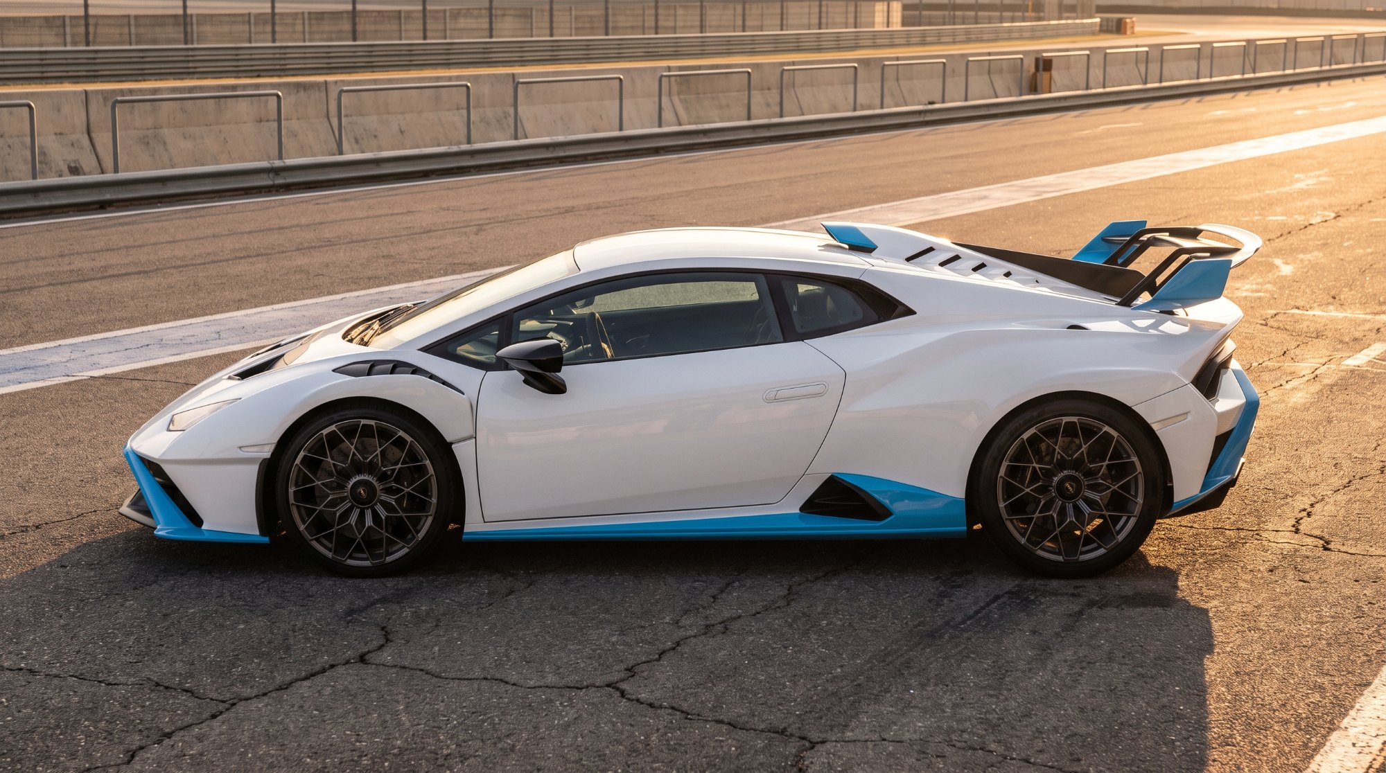 White and light-blue Lamborghini Huracán STO in side profile in a race-circuit pit lane, showing its roof scoop and large rear wing